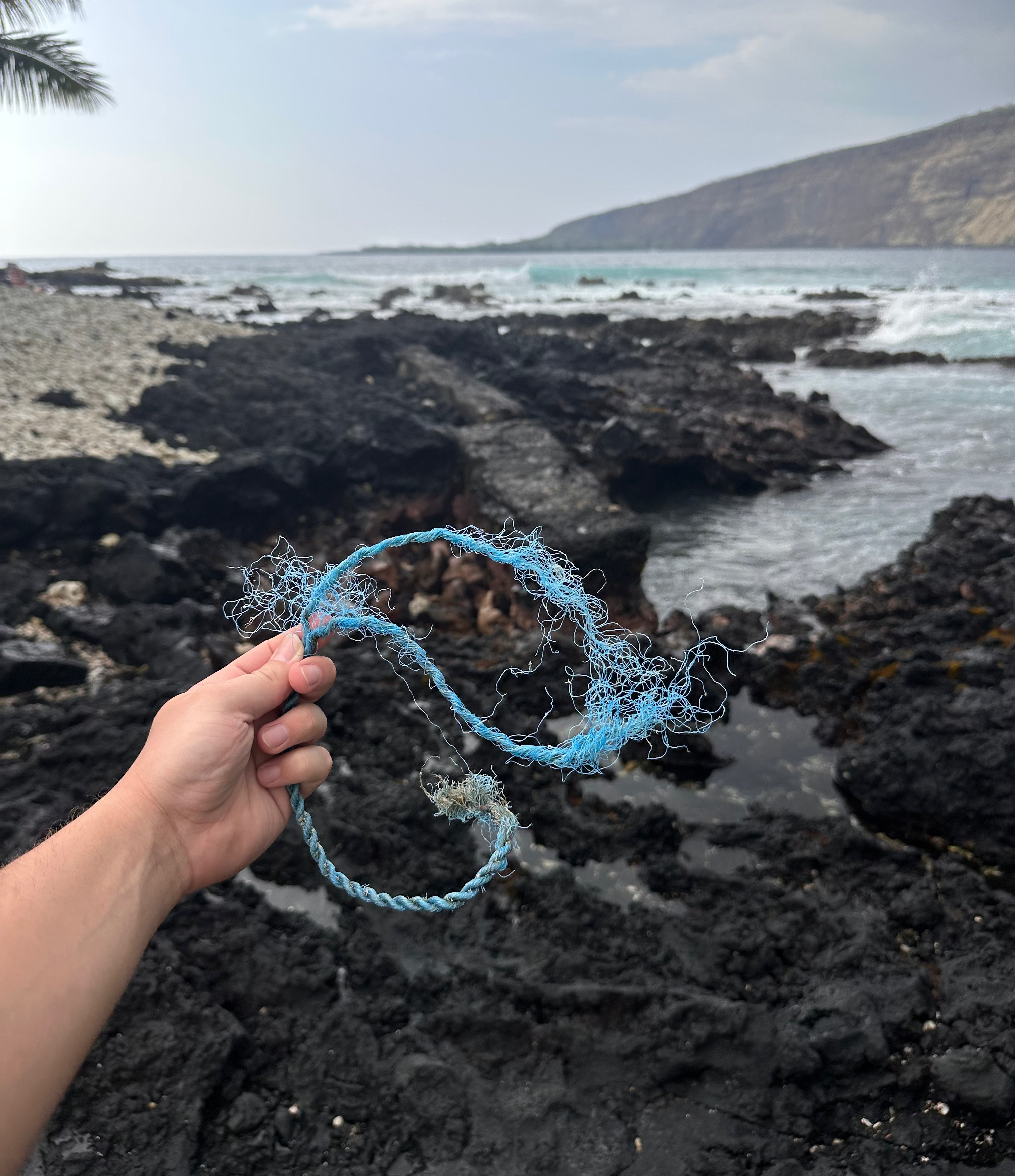 Hand holding trash on a rocky beach with ocean and mountains in the background in Hawaii