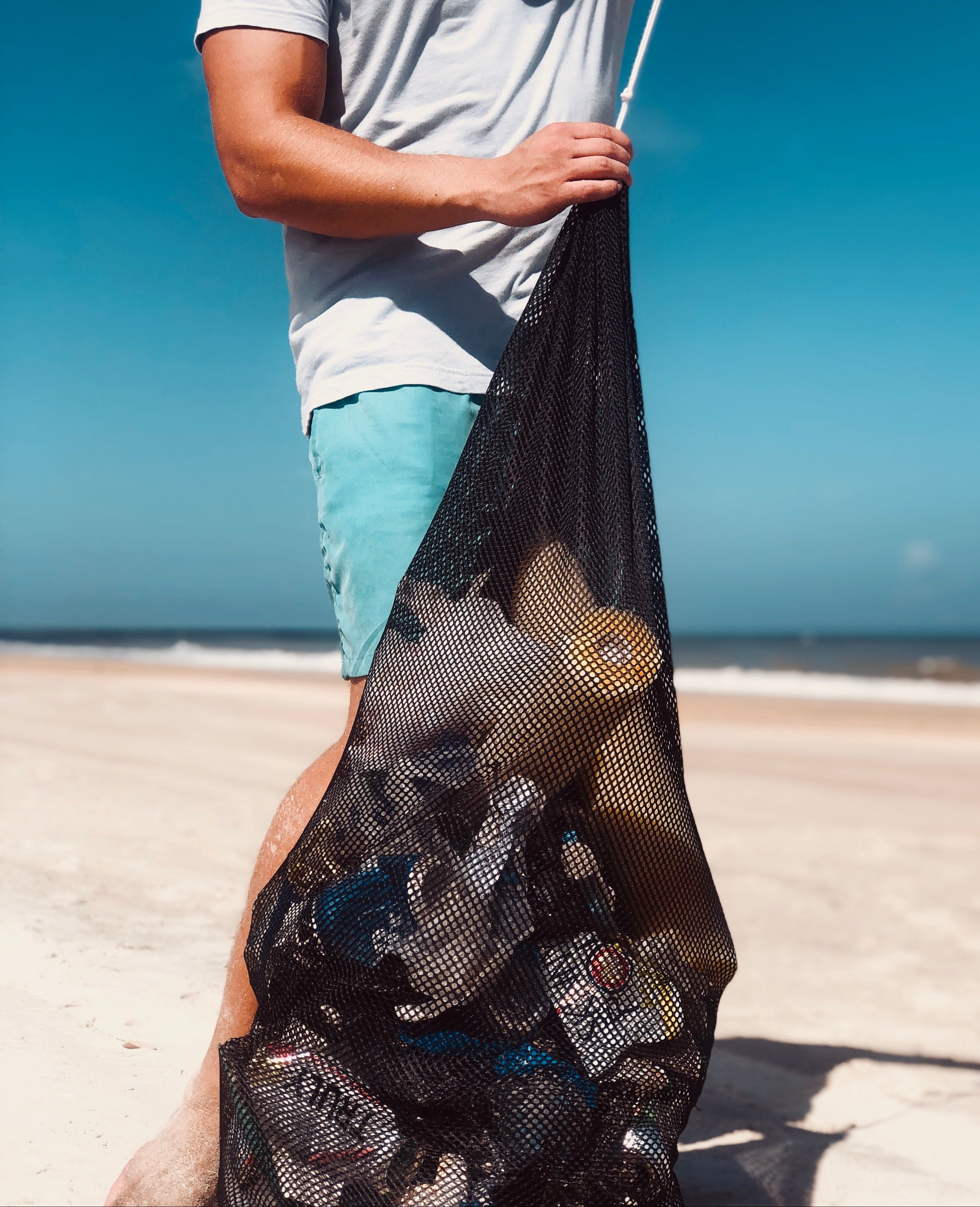 Person holding a black mesh bag on a beach cleanup with a clear blue sky.