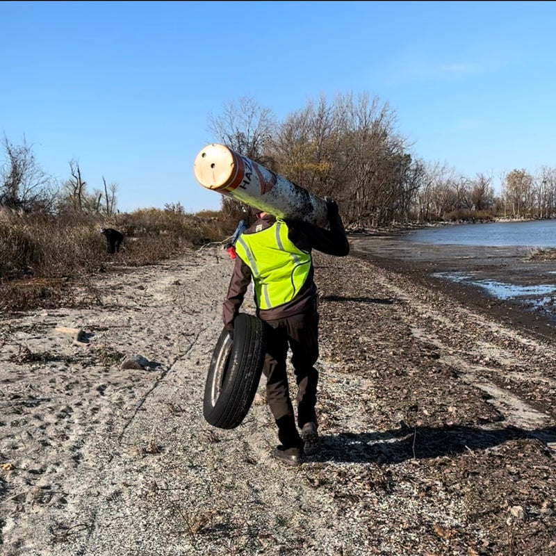 Person in a yellow safety vest carrying beach trash on a beach near water