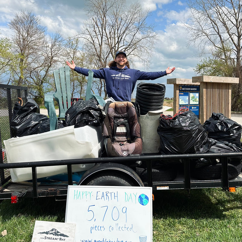Person with arms outstretched next to a trailer filled with trash and a 'Happy Earth Day' sign.