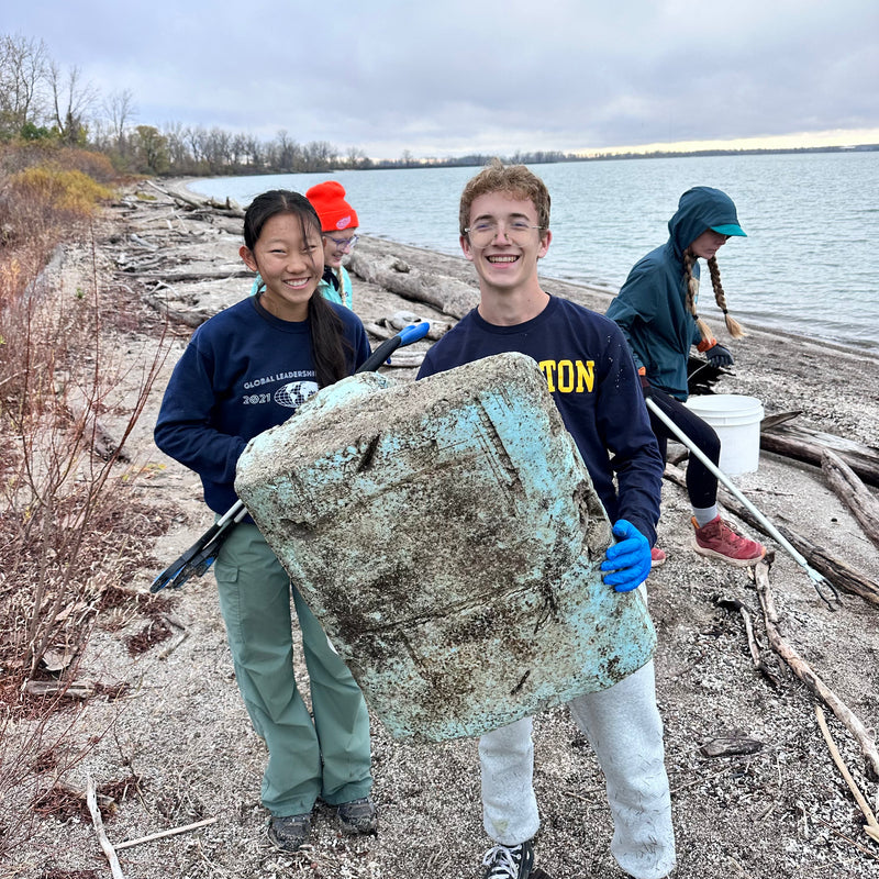 Two people holding a large piece of marine debris on a beach with water and sky in the background.